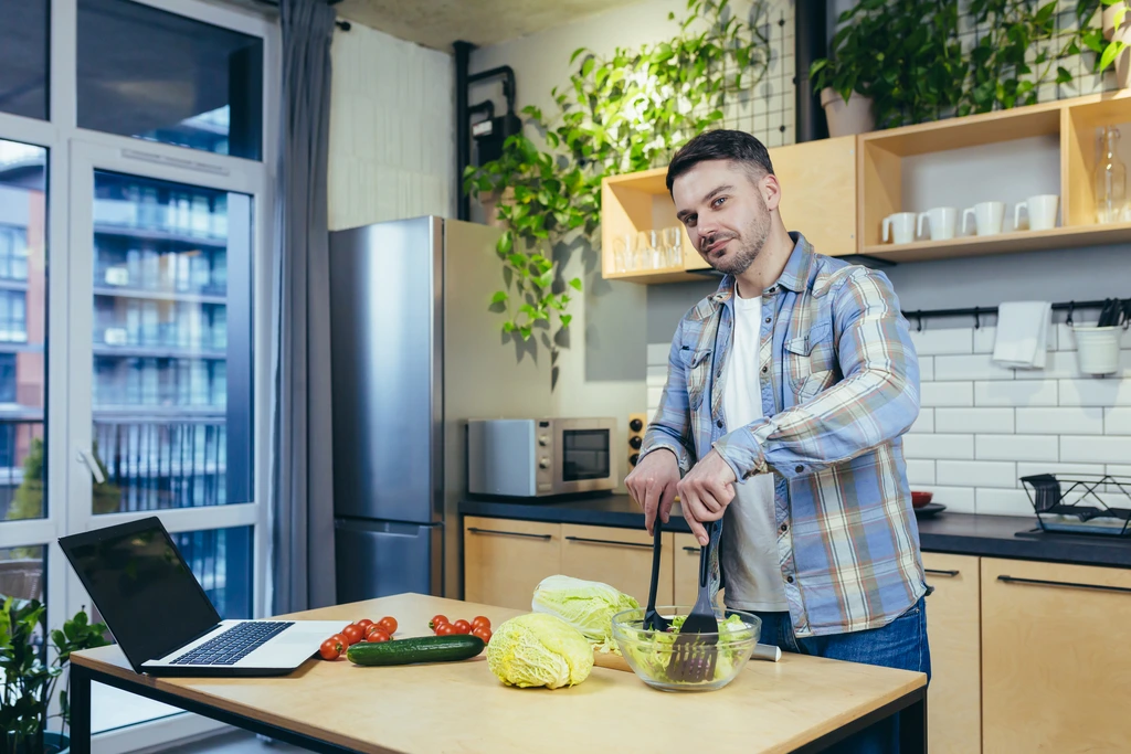 Mike Carter from Delish Symphony mixing a salad in a bowl, with his laptop open on the kitchen counter beside him.