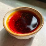 A close-up overhead shot of a small ceramic bowl filled with a rich, dark red and amber-colored dumpling sauce, speckled with chili flakes.