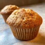 Close-up of two golden-brown banana protein muffins in paper liners, with one in focus.