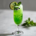 A vibrant green Beetlejuice Prosecco cocktail in a champagne flute, garnished with lime and mint, and decorated with googly eyes.