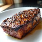 A close-up of a perfectly cooked Blackened Tuna Steak, seasoned with spices and resting in its juices on a white plate.