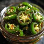 A close-up, overhead view of bright green candied jalapenos glistening in a glass jar, with visible seeds and red pepper flakes.