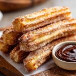 A stack of golden-brown churros coated in cinnamon sugar, served with a small bowl of rich chocolate dipping sauce.