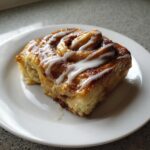 A single slice of cinnamon roll casserole with white icing drizzled on top, served on a white plate.