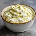 Close-up of creamy garlic aioli with herbs and oil drizzled on top in a rustic bowl.