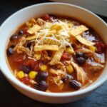 A close-up of a white bowl filled with Crockpot Chicken Enchilada Soup, topped with shredded cheese and tortilla strips.