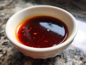 Close-up of a small white bowl filled with rich, dark red dumpling sauce with visible chili flakes.