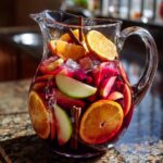 A clear glass pitcher filled with vibrant red fall sangria, loaded with orange slices, apple wedges, cranberries, and cinnamon sticks.