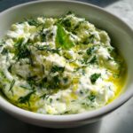 A close-up of creamy Tzatziki sauce topped with fresh dill, parsley, mint, and lemon zest.