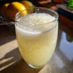 A close-up of a glass filled with frothy Frozen Creamy Lemonade, condensation on the glass, with lemons in the background.