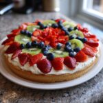 A vibrant fruit pizza sugar cookie topped with cream cheese frosting, strawberries, blueberries, and kiwi slices.