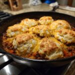 A close-up of a hearty ground beef casserole topped with golden-brown biscuits and melted cheddar cheese in a cast-iron skillet.