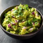 A close-up of a dark bowl filled with chunky, vibrant Homemade Guacamole, topped with fresh cilantro and red onion.
