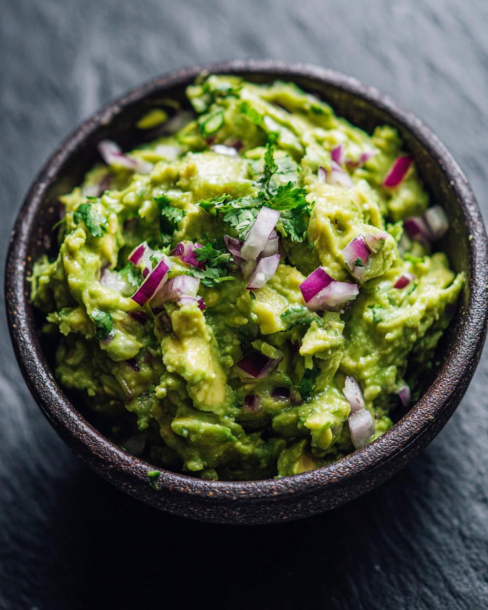 A close-up of vibrant, chunky homemade guacamole in a dark bowl, topped with red onion and cilantro.