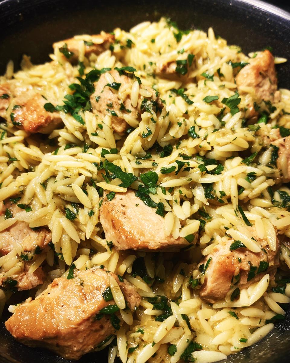 Close-up of a pan filled with lemon chicken orzo, featuring tender chicken pieces and orzo pasta mixed with fresh parsley.