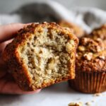 A hand holds a moist banana nut muffin, showing its fluffy interior with chunks of banana and walnuts.