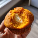 A hand holds a Starbucks pumpkin cream cheese muffin with a bite taken out, revealing the creamy center.
