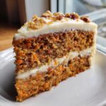 A tall, moist slice of carrot cake recipe showing visible shredded carrots, layered with cream cheese frosting and topped with walnuts.
