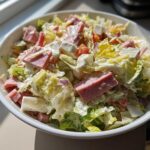 A close-up, bright photo of a hearty grinder salad featuring chopped lettuce, pink ham cubes, white cheese, and creamy dressing.
