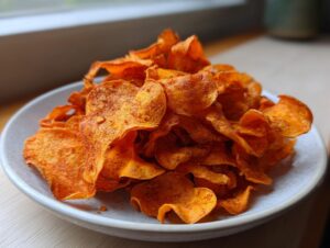 A close-up of a large serving of seasoned, crispy sweet potato chips piled high on a light gray plate.
