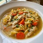 A close-up, appetizing view of homemade chicken noodle soup featuring shredded chicken, egg noodles, and sliced carrots, garnished with parsley.