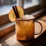 A refreshing apple cider moscow mule cocktail served over crushed ice in a copper mug, garnished with an apple slice and cinnamon sticks.