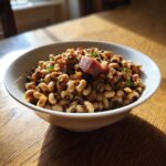 A white bowl filled with a savory black eyed peas recipe, topped with a piece of ham or smoked meat.