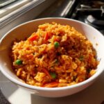 A close-up of a white bowl filled with vibrant orange Mexican rice mixed with bright green peas and sliced carrots.