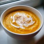 Close-up of a bowl of vibrant orange butternut squash soup, topped with a swirl of cream and a sprinkle of spice.