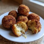 A plate of golden brown, crispy rice balls, with one cut open showing a gooey, melted cheese pull inside.