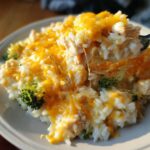 A spoonful of cheesy chicken and rice casserole being lifted, showing melted cheddar cheese strings and pieces of broccoli.