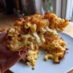 A hand holding a massive, cheesy serving of baked mac and cheese with a crispy topping, showing long cheese pulls.