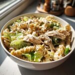 A close-up of a bowl filled with chicken caesar pasta salad, featuring rotini pasta, shredded chicken, and romaine lettuce tossed in creamy dressing.