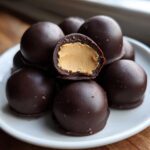 A stack of dark chocolate covered peanut butter balls, with one cut in half showing the creamy peanut butter center.