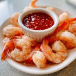 A plate of chilled, peeled shrimp arranged around a small white ramekin of spicy red shrimp cocktail sauce.