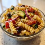A close-up shot of a glass bowl filled with vibrant greek pasta salad featuring rotini pasta, cucumbers, tomatoes, olives, and red onion.