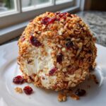 A creamy, round cranberry pecan cheese ball coated heavily in chopped nuts and dried cranberries, resting on a white plate.