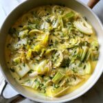 Close-up overhead view of creamy braised leeks cooked in a light-colored skillet, garnished with herbs.