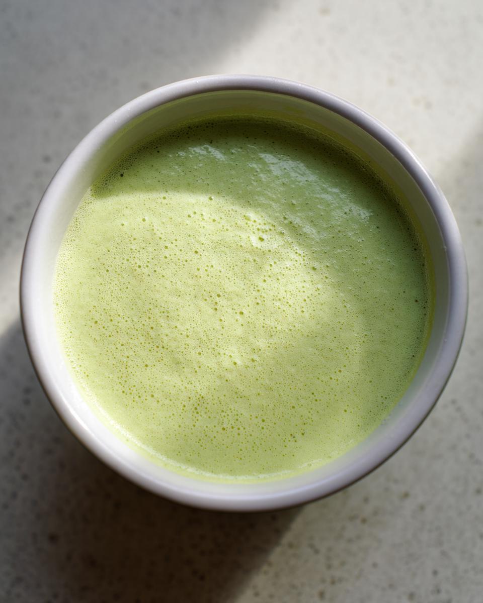 Overhead view of creamy, light green celery soup with a slight froth in a white bowl.