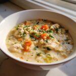 A white bowl filled with creamy chicken and rice soup, featuring chunks of chicken, bright orange carrots, and fresh parsley.