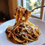 A fork lifting a generous portion of creamy french onion pasta topped with grated cheese from a white bowl.