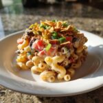 A mound of creamy taco pasta salad featuring elbow macaroni, tomatoes, olives, and topped with crushed tortilla chips and green onions.