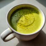 Close-up of a vibrant green, creamy zucchini soup in a white mug, topped with a swirl of olive oil and black pepper.