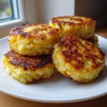 A stack of four golden brown, crispy mashed potato cakes resting on a white plate near a window.