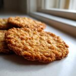 Close-up of several golden brown, crispy oatmeal lace cookies resting on a light countertop near a window.