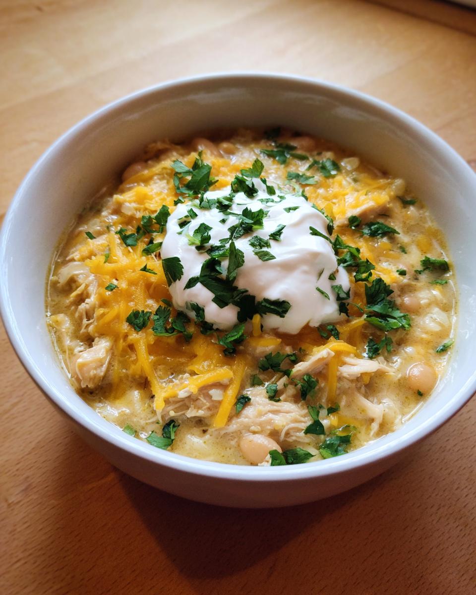 A close-up of a bowl of creamy crockpot white chicken chili topped with shredded cheese, sour cream, and fresh parsley.