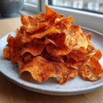 A close-up of a mound of crispy, seasoned sweet potato chips piled high on a light grey plate.