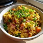 Close-up of a white bowl filled with bright yellow cauliflower fried rice, mixed with egg, carrots, and topped with fresh green scallions.