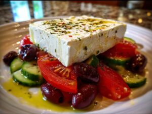 A large block of feta cheese seasoned with herbs sits atop sliced tomatoes, cucumbers, and Kalamata olives in a fresh greek salad.