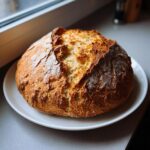 A golden-brown, rustic loaf of freshly baked soda bread with a cracked crust, resting on a white plate.